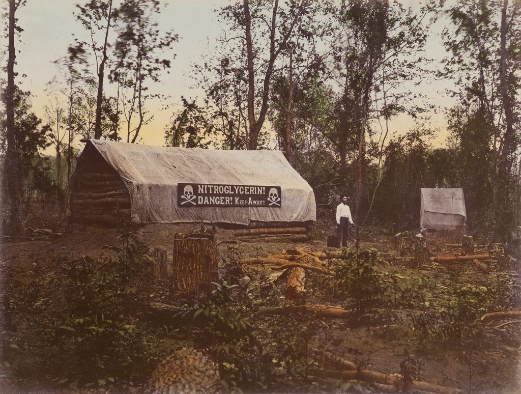 A hand-colored photograph of red river raft which features the brush and swamp of Louisiana with fallen trees surrounding a log cabin with a tarp over the roof that reads NITROGLYCERIN! DANGER! KEEP AWAY! with a man standing in front.