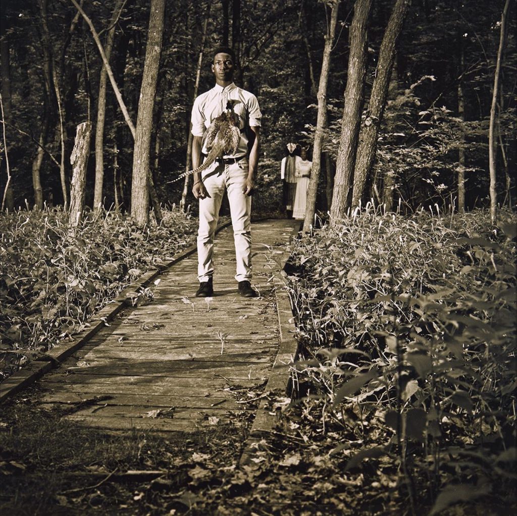 Photograph of a young black man with a pheasant attached to his chest by Allison Janae Hamilton. 