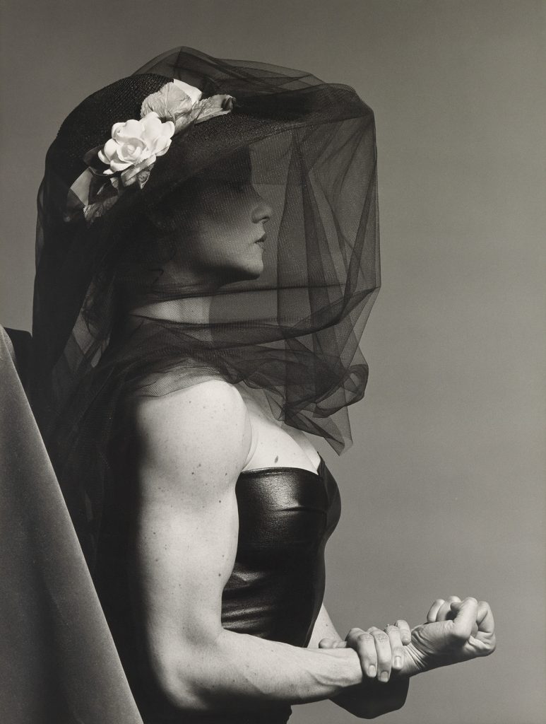 Black and white photograph of a veiled Lisa Lyon flexing her right arm by Robert Mapplethorpe. 