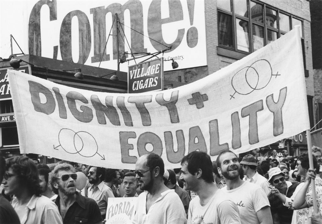 Photograph of parade goers at a NYC Pride parade in front of Village Cigars with a sign that reads "Dignity + Equality."