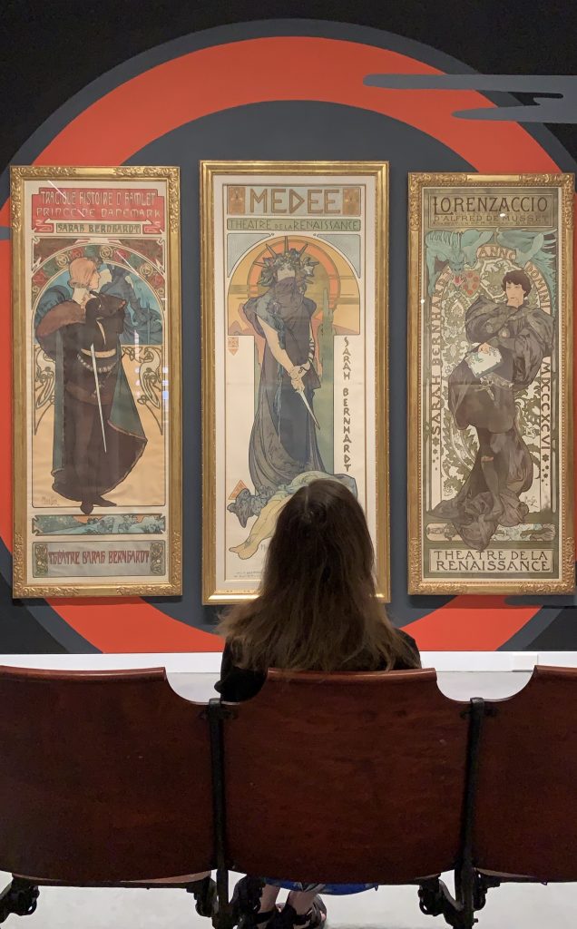 Lauren Goldberg sits in old theater chairs at the Poster House museum's Alphonse Mucha exhibition.