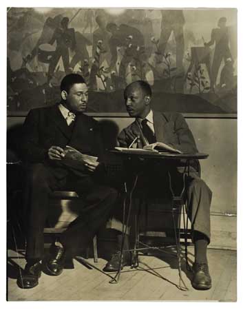 Black and white photograph of Frederick O'Neal & Abram Hill seated in front of Aaron Douglas's mural at the Schomburg Center for Research in Black Culture, circa 1940-45.